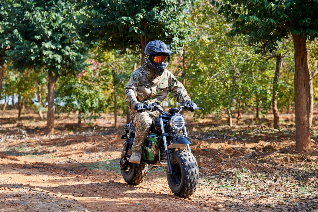 A person in camouflage gear and helmet rides a MotoTec 60v 1500w Electric Powered Mini Bike Lithium Black on a dirt path surrounded by trees.