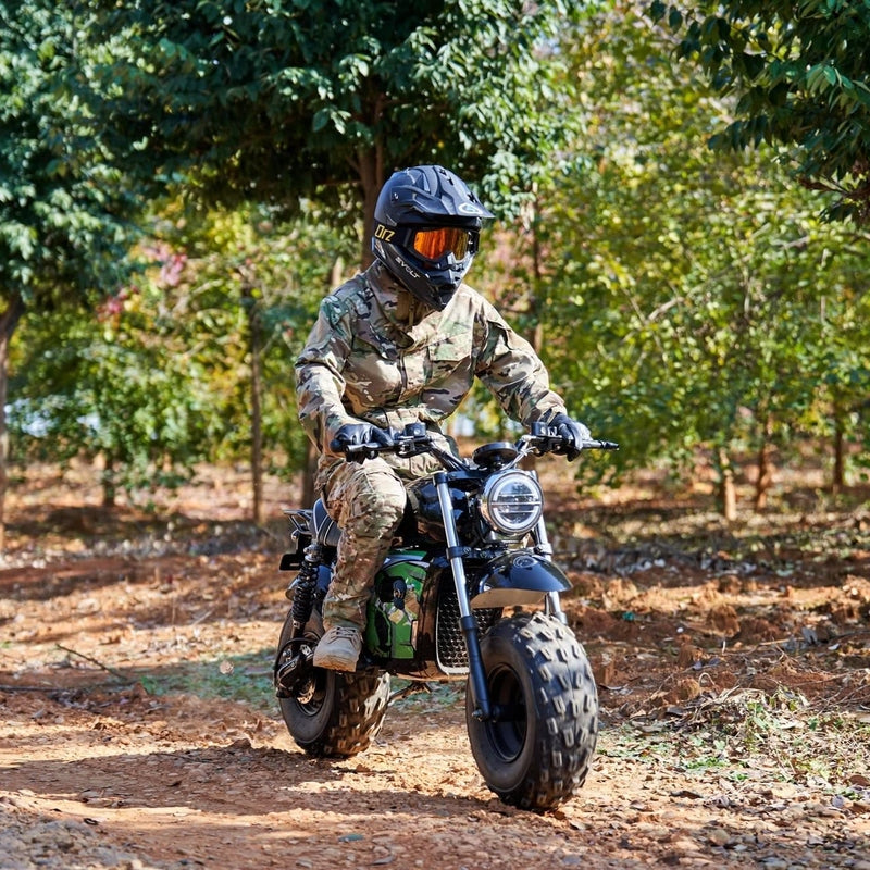 A person in camouflage gear and helmet rides a MotoTec 60v 1500w Electric Powered Mini Bike Lithium Black on a dirt path surrounded by trees.