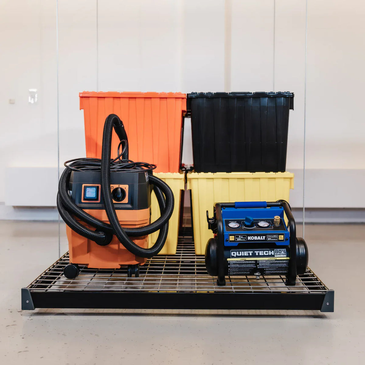 Black-framed Auxx-Lift platform at floor level holding a shop vacuum, air compressor, and totes