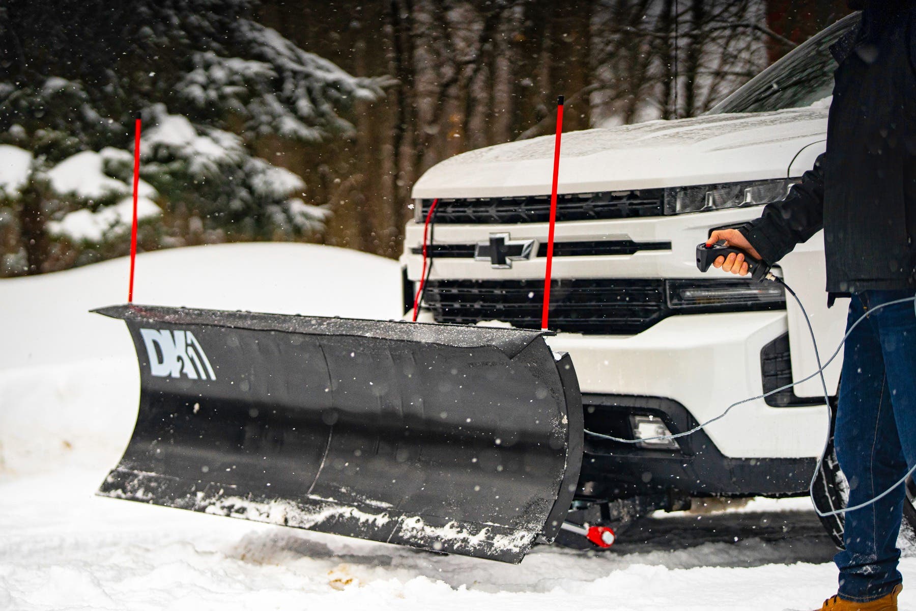Operator angling the DK2 Everest plow with the wired handheld remote beside a white Chevrolet pickup