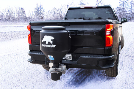 SnowBear salt spreader mounted in the rear hitch receiver of a pickup truck on a snowy road