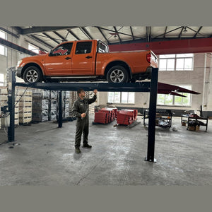 Technician working beneath an orange pickup raised on the four-post lift in a shop